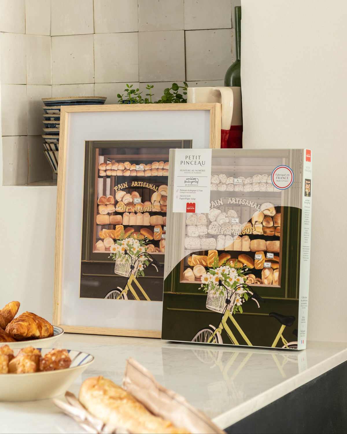 A framed and boxed art print of a Parisian bakery storefront with breads and pastries, displayed on a kitchen counter next to a bowl of croissants and a baguette.
